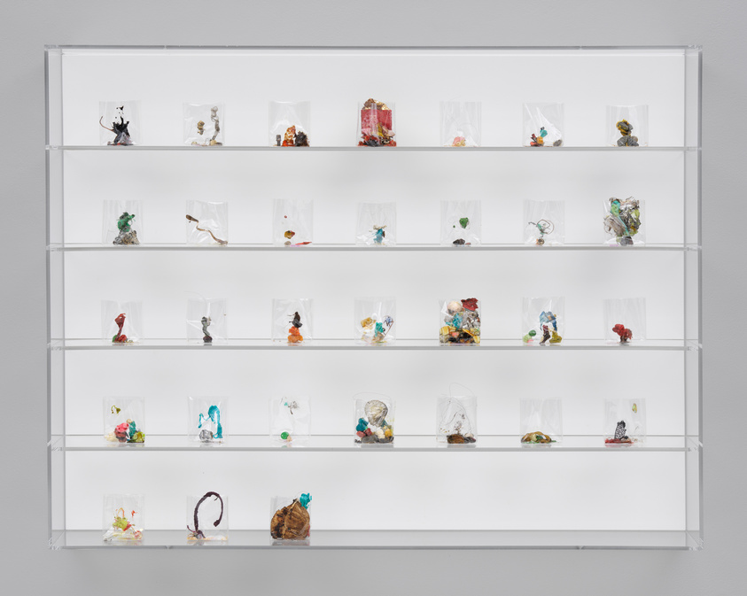 Clear display case with four shelves, each holding small plastic bags containing colorful mixed objects or materials.