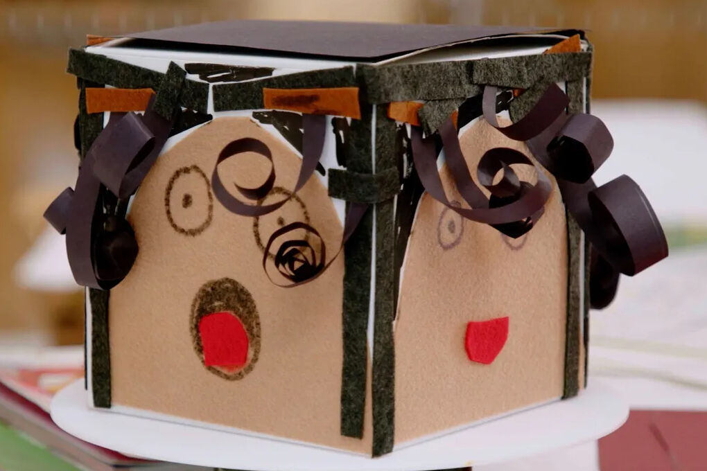 Two hands holding a box each side showing a self portrait made of paper, curled ribbon, and drawings.