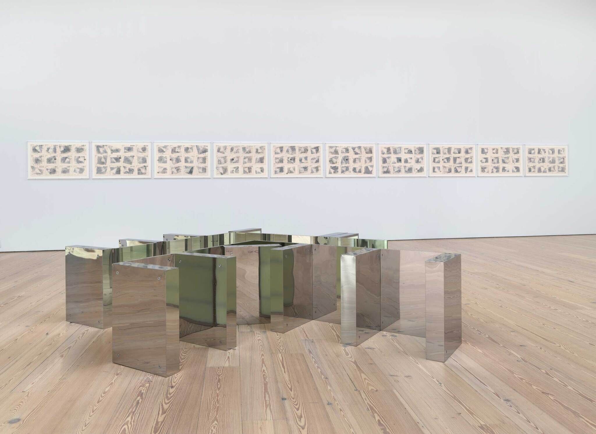 Reflective sculpture on wooden floor with framed artworks on the wall in a gallery setting.