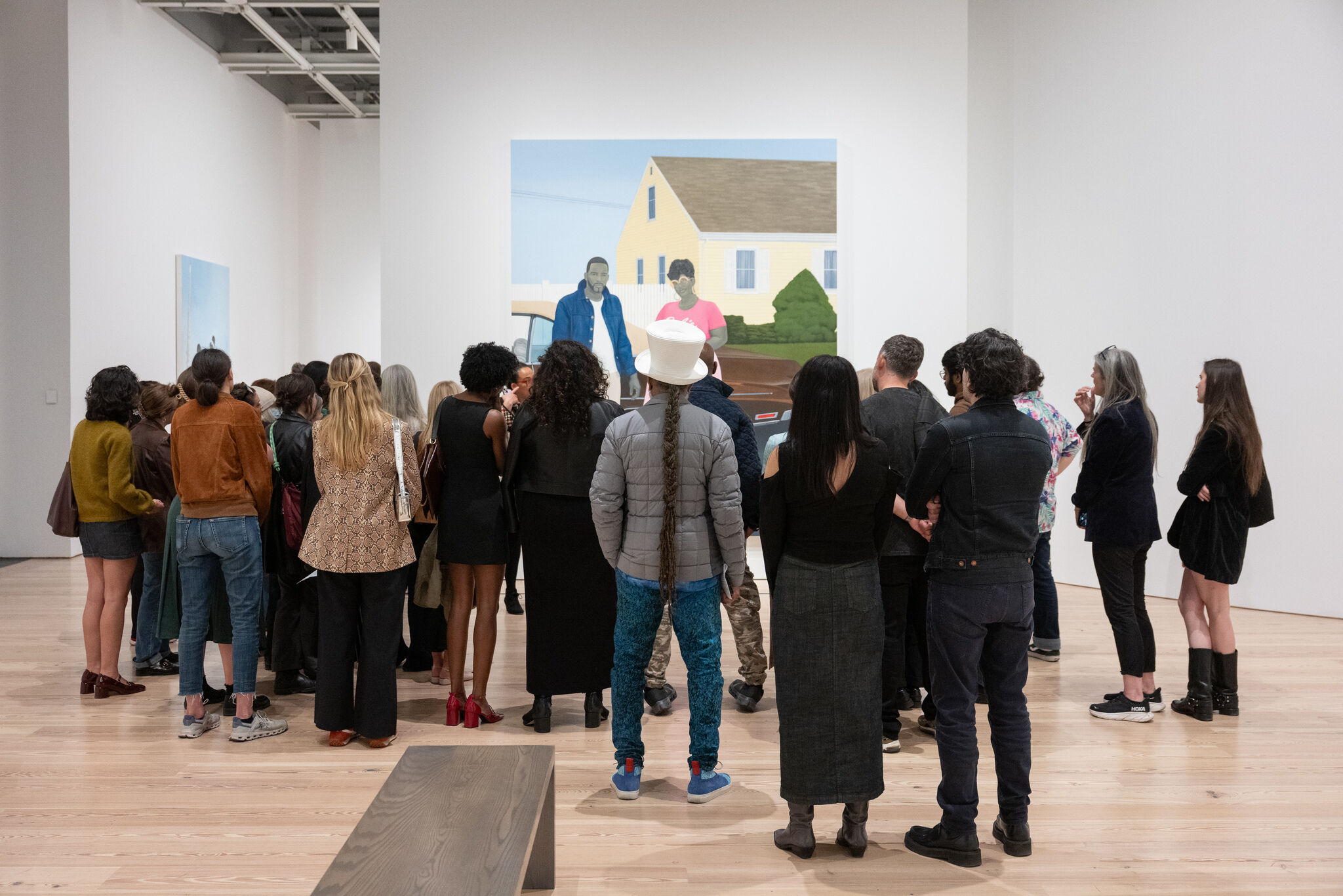 A group of people stands closely together in an art gallery, looking at a large painting on the wall.
